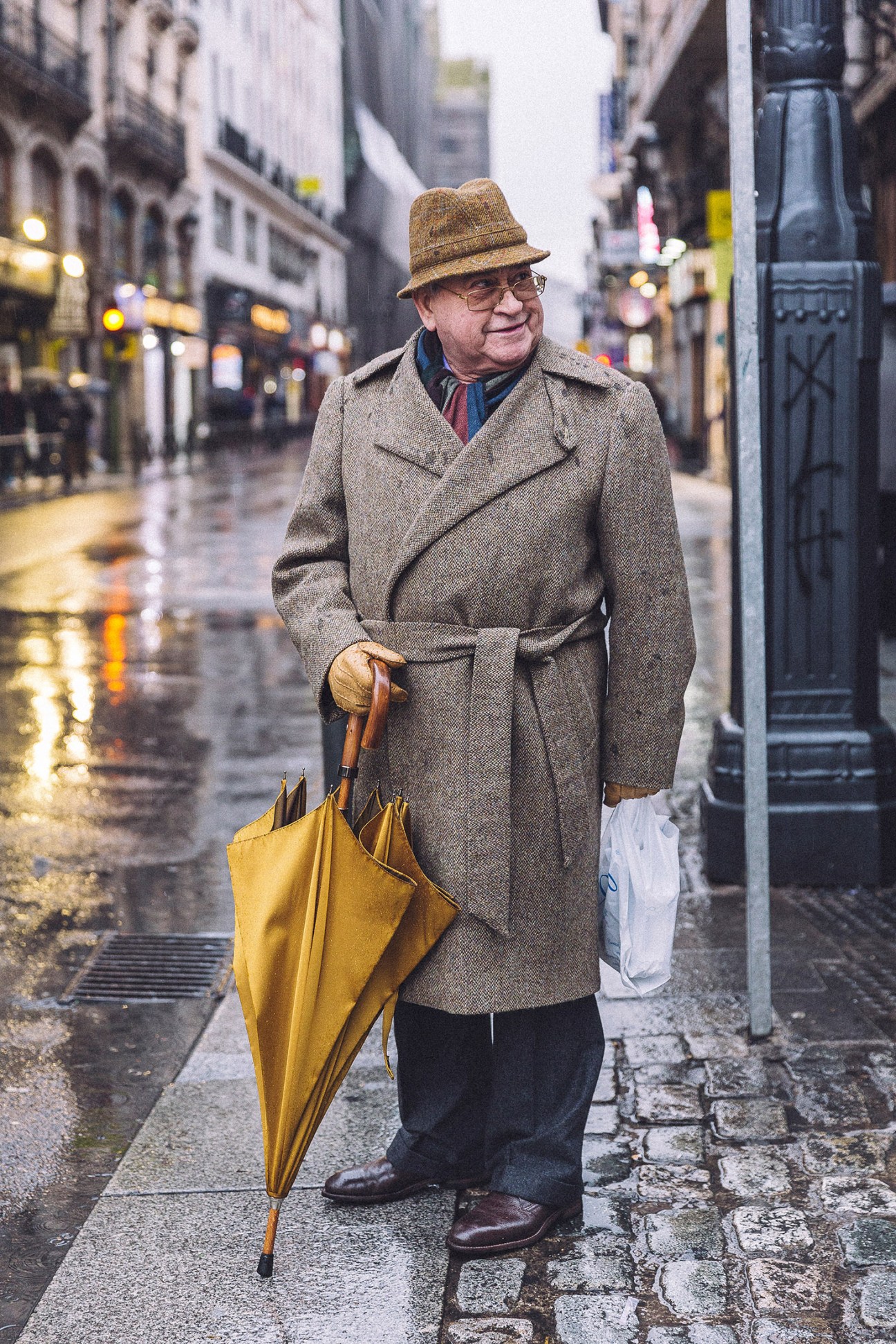 Elderly man in brown coat and fedora holding yellow umbrella on rainy Madrid street.