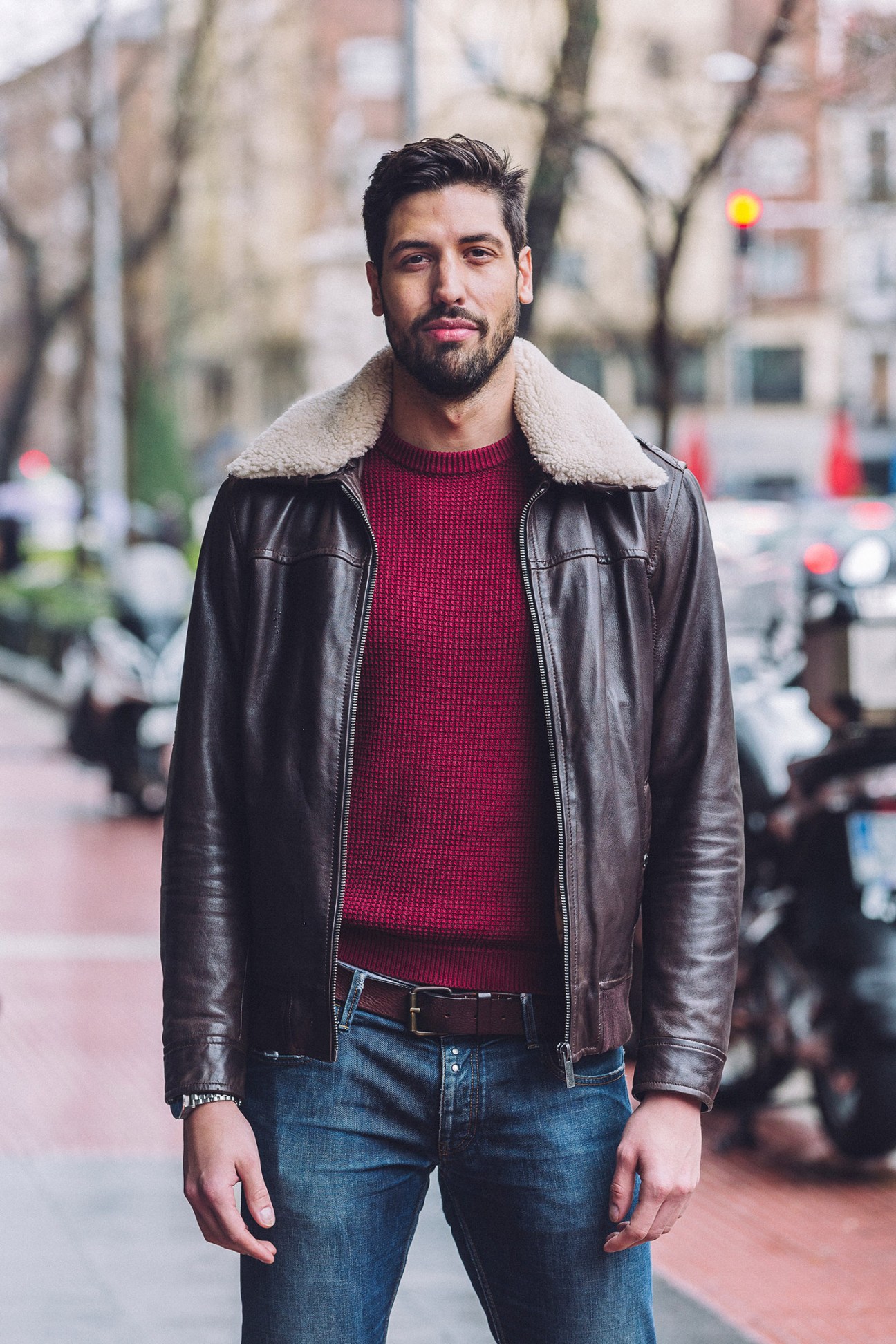 Man with dark hair and beard wearing black leather jacket with sherpa collar and burgundy knit sweater on Madrid street.