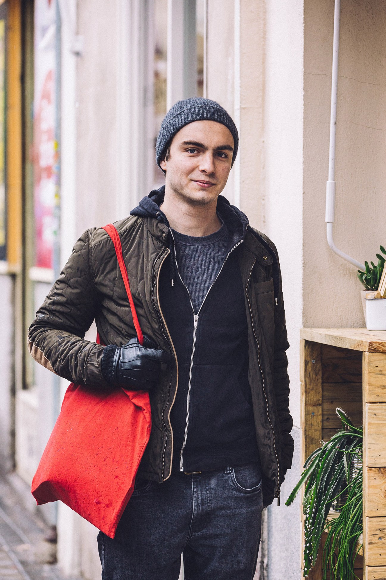 Man wearing grey beanie, black puffer jacket and red tote bag standing on a Madrid street.