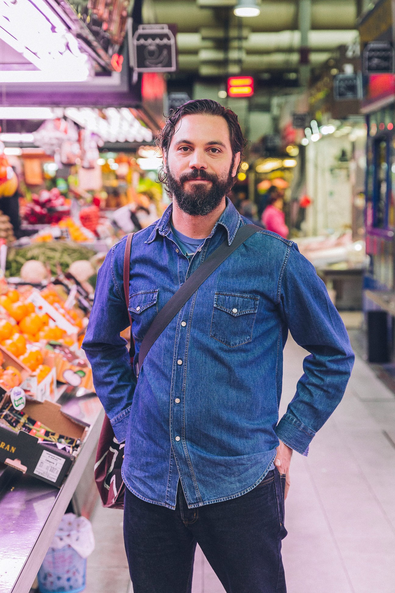 Man in denim shirt standing in a colorful market with produce displays in the background.