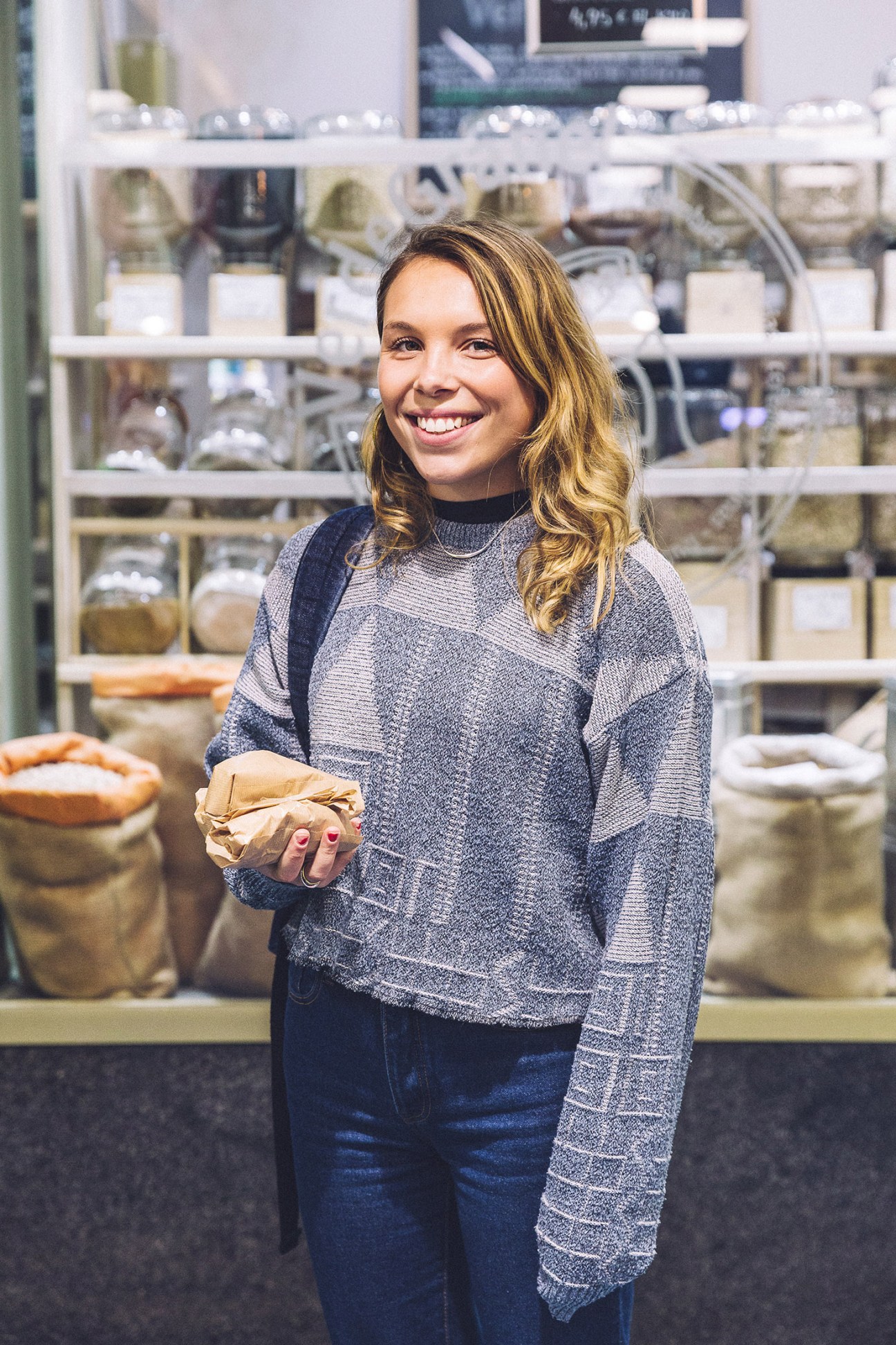 Young woman holding a wrapped sandwich in a food market with shelves of products behind her.