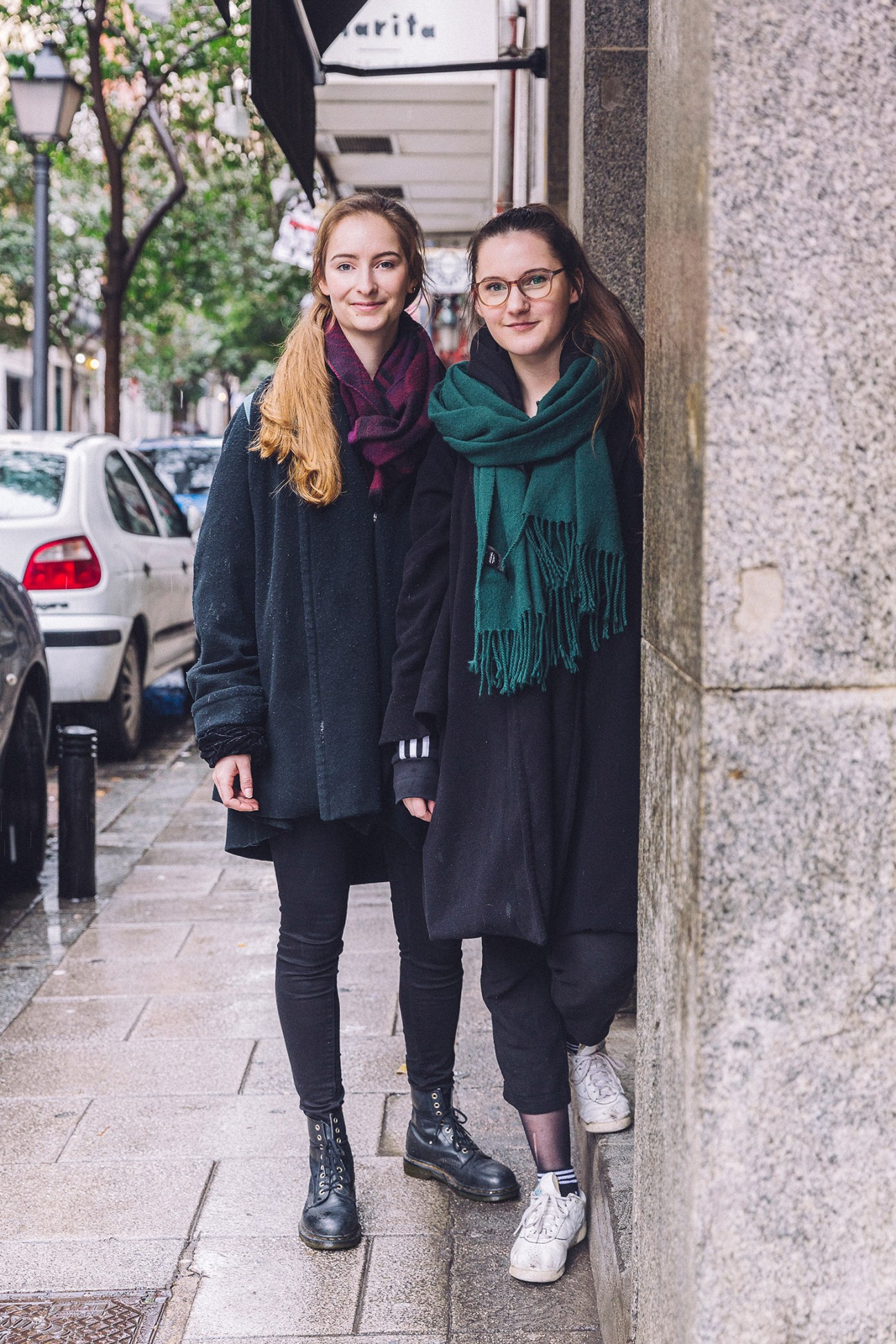 Two young women stand smiling on a Madrid street lined with blooming trees and parked cars.