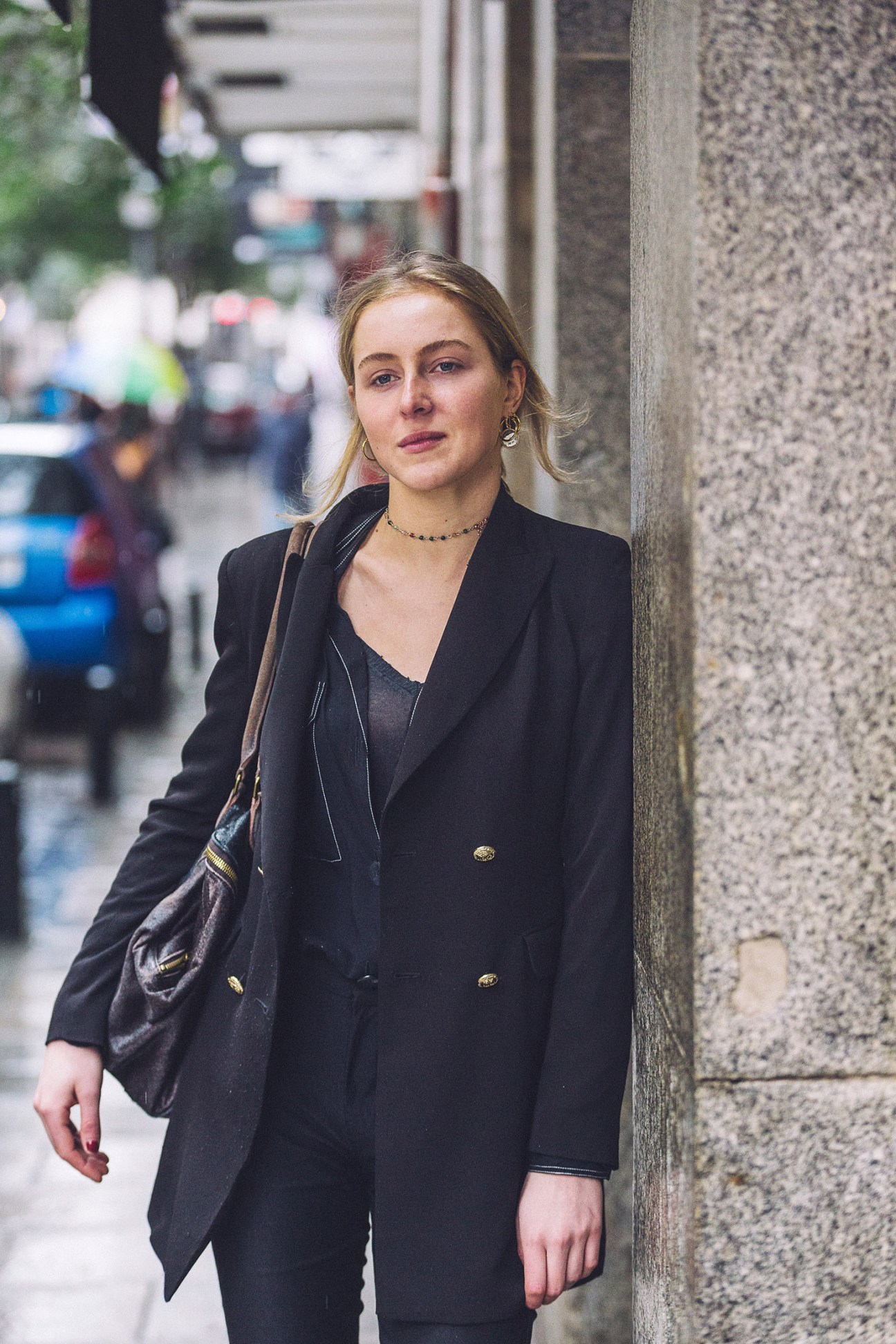 Woman in black blazer and beaded necklace standing against textured grey wall on Madrid street