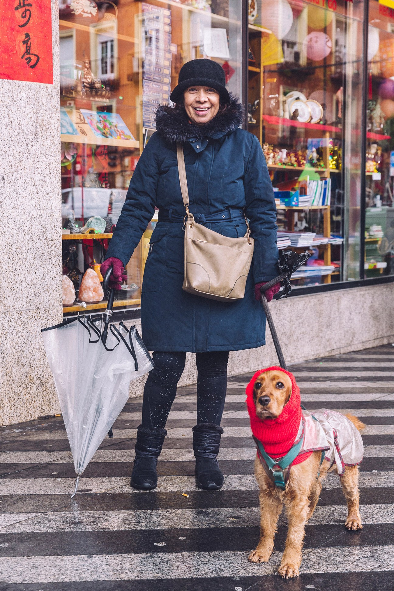 Woman in navy coat and black hat walks dog wearing red knit sweater outside storefront.
