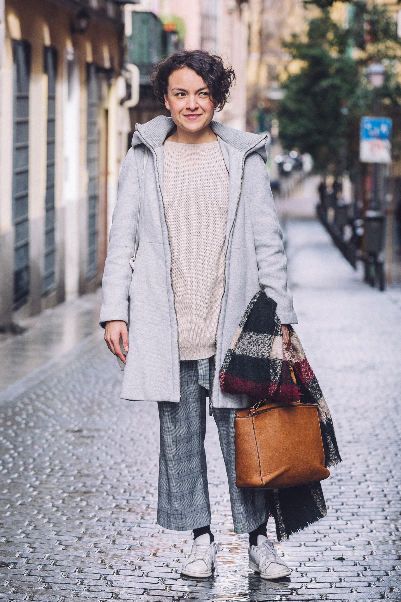 Woman with short dark hair wearing a light gray coat and checkered pants, carrying a patterned scarf and tan leather bag on a European street.