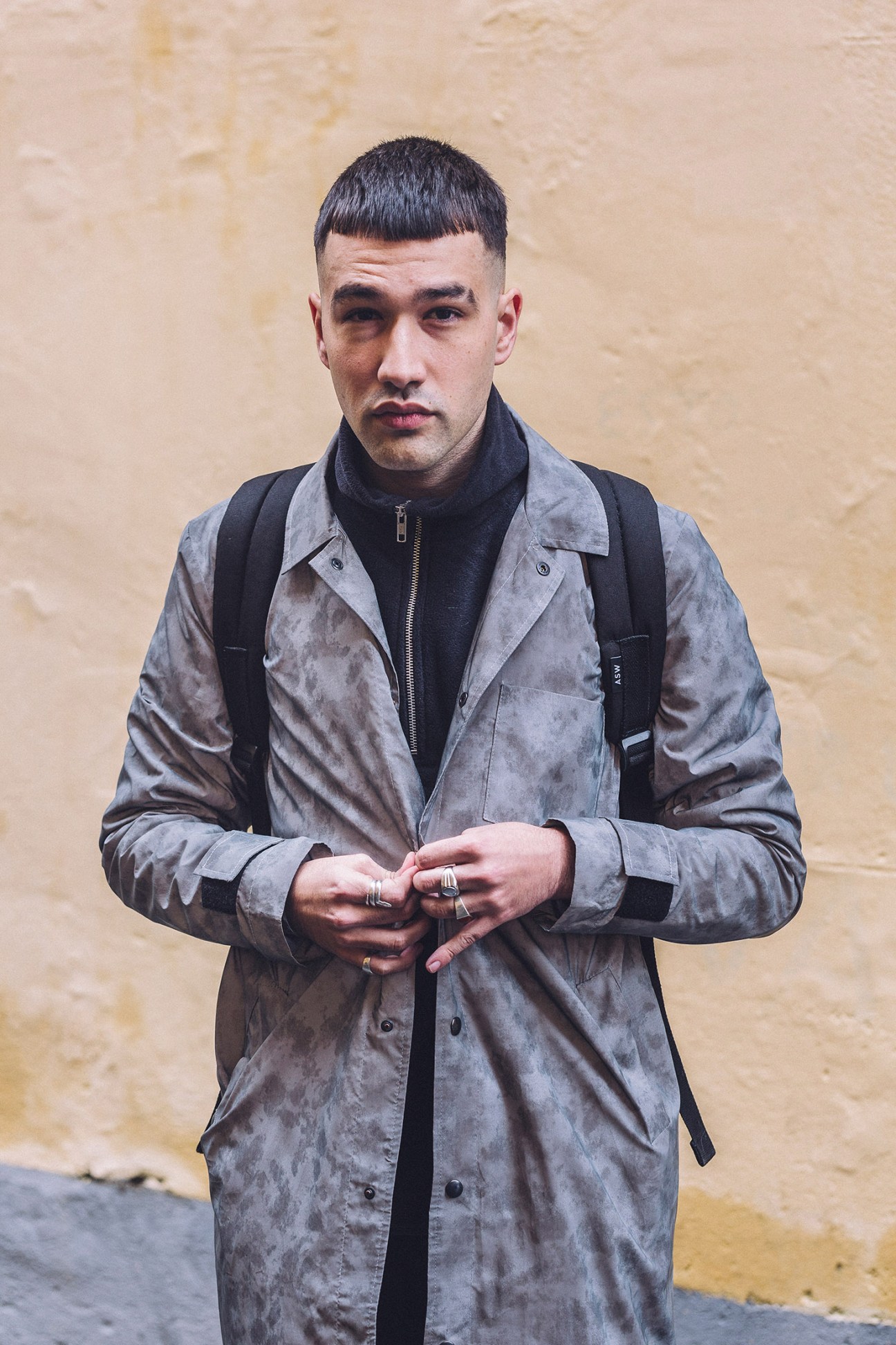 Man in grey suede jacket and black turtleneck standing against beige wall