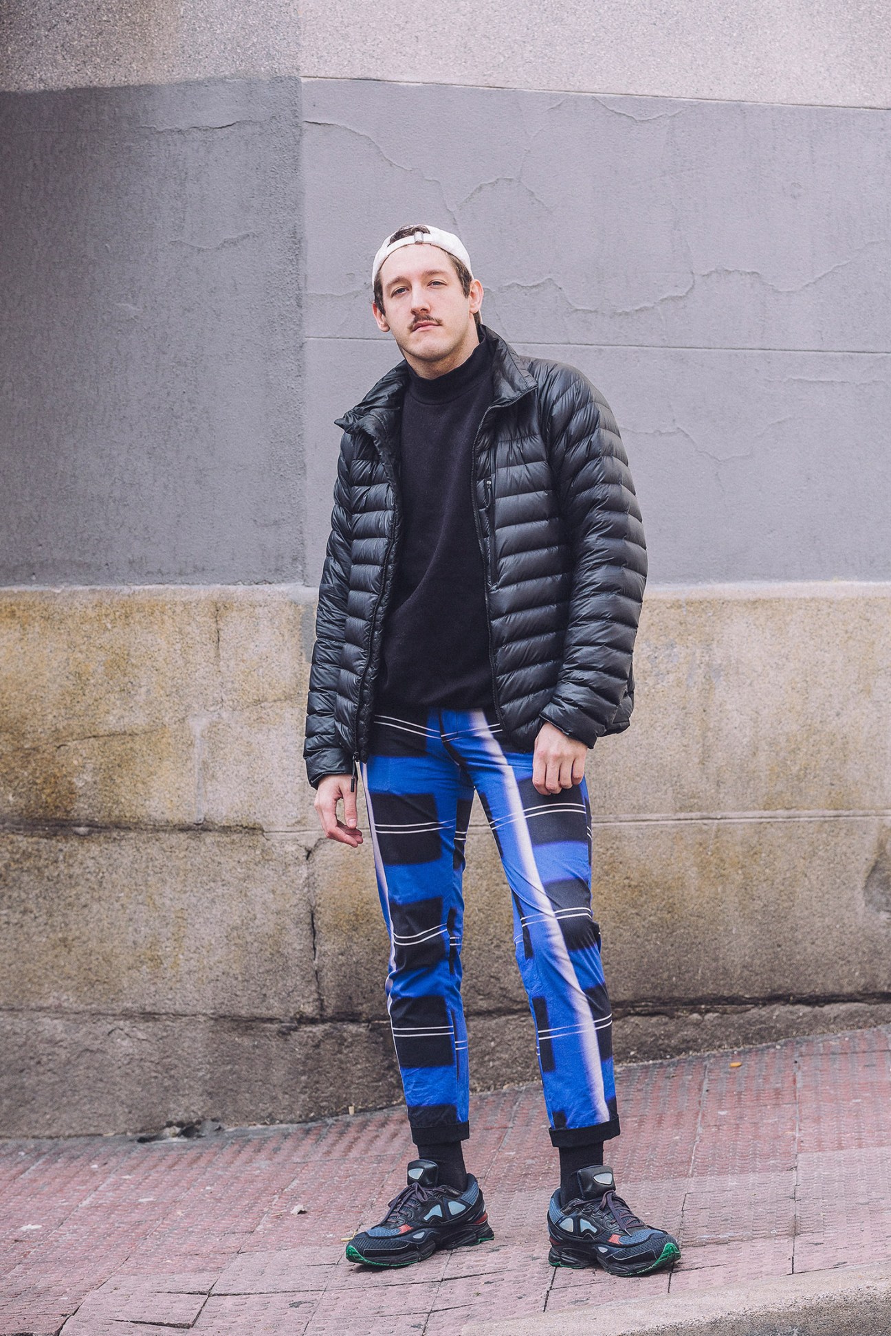 Young man wearing white headband, black puffer jacket, and blue geometric patterned pants stands against urban concrete wall.