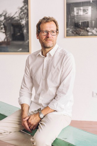Man in white shirt and glasses sitting on bench with framed photographs on wall behind him.