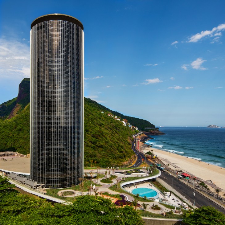 Cylindrical modernist hotel tower overlooking a coastal beach with green hillside and ocean beyond.