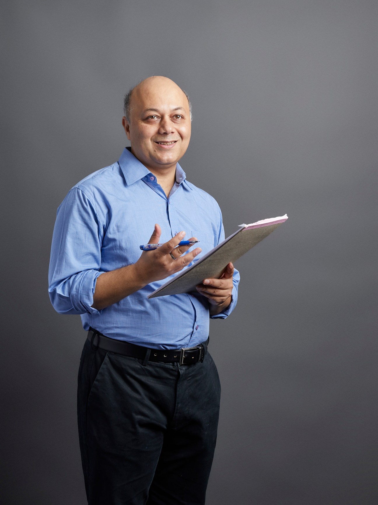 Man in blue shirt holding tablet and pen, smiling at camera against gray background.