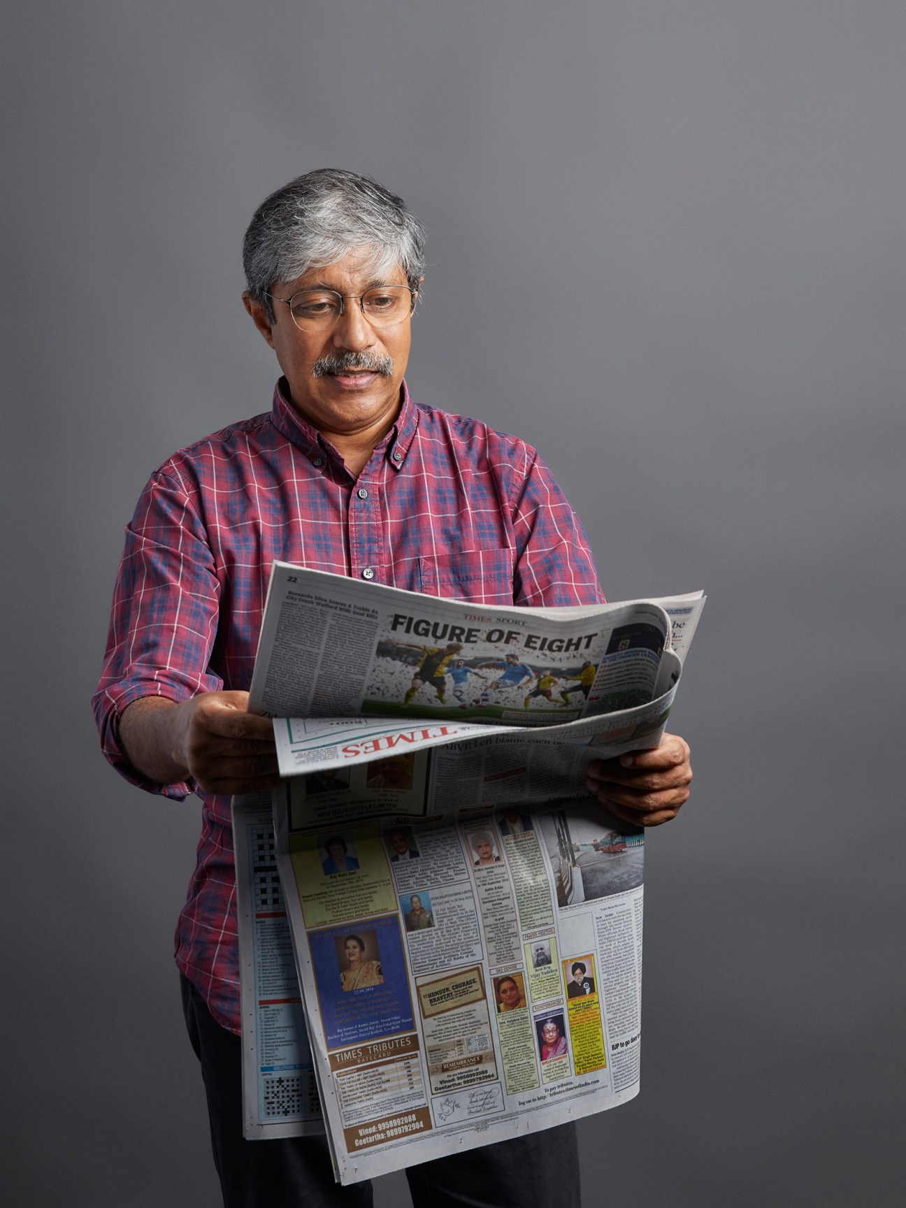 Man in plaid shirt reading Times of India newspaper against gray background.