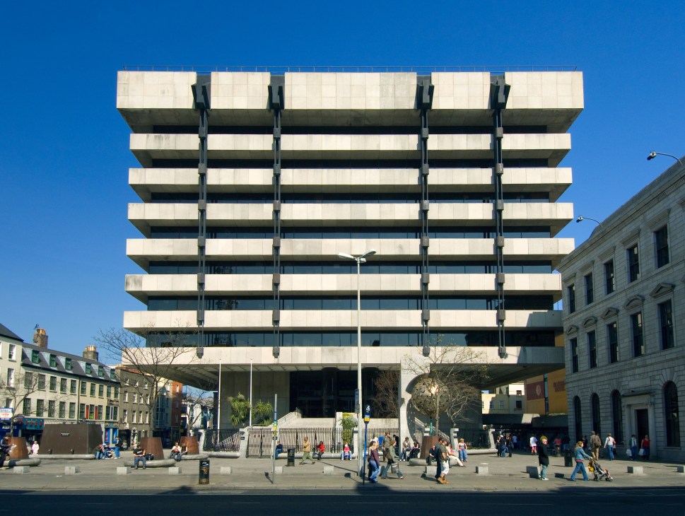 Dublin's Central Bank building, a brutalist concrete structure with horizontal bands of stone and glass against a clear blue sky.