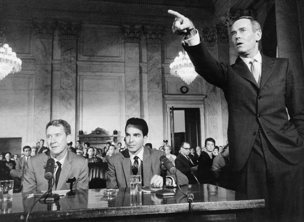 Black and white still from the 1962 film Advise and Consent showing men in suits at a formal hearing.
