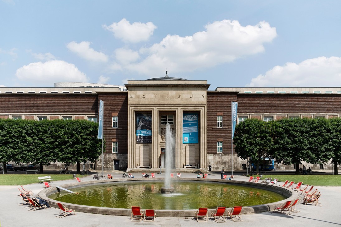 Neoclassical museum building with brick wings, central portico with fountain and red deck chairs in foreground.