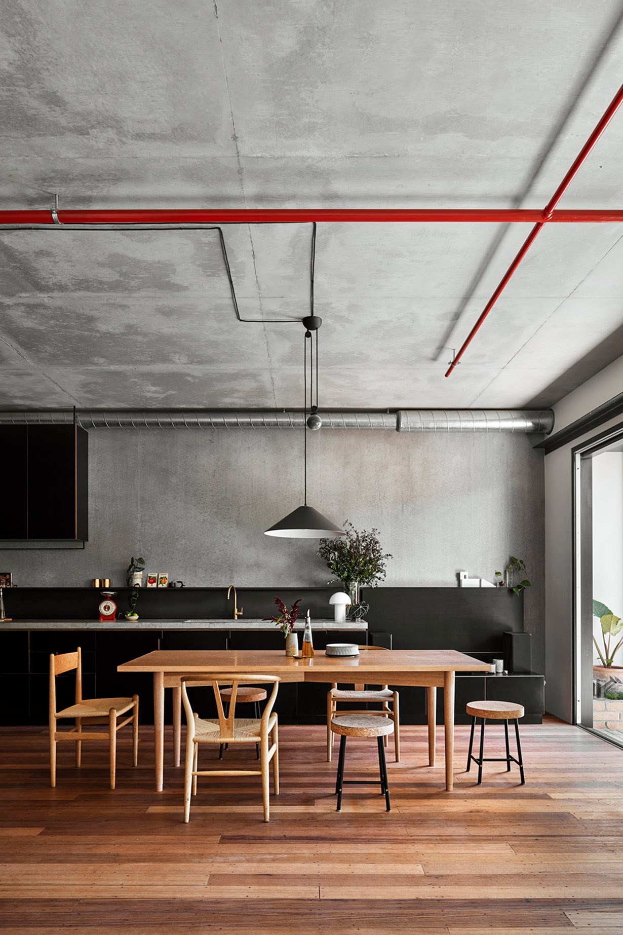 Modern loft interior with exposed concrete ceiling, red pipes, wooden dining table, and minimalist kitchen.