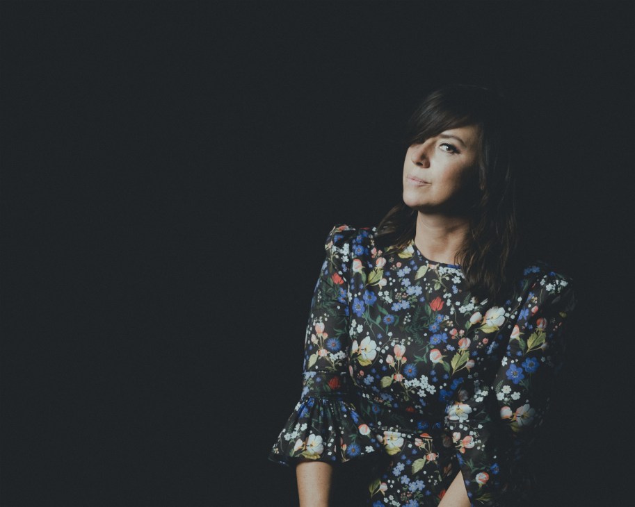 Woman with dark hair wearing a black floral print dress against a dark background