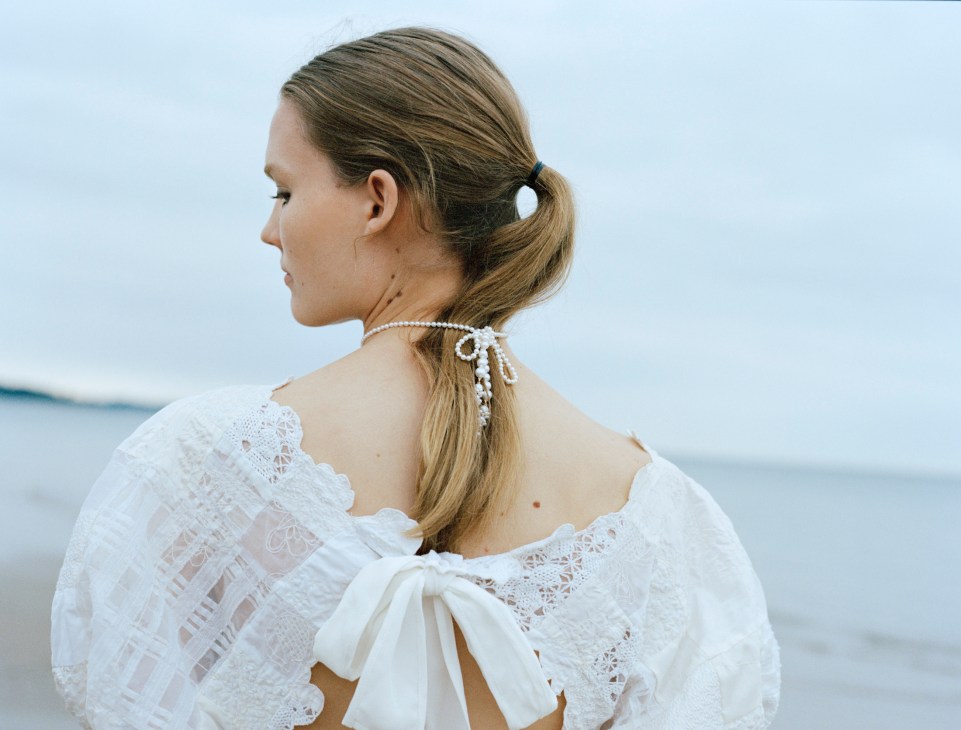 Woman in white lace dress with pearl hair accessory looking toward ocean horizon.
