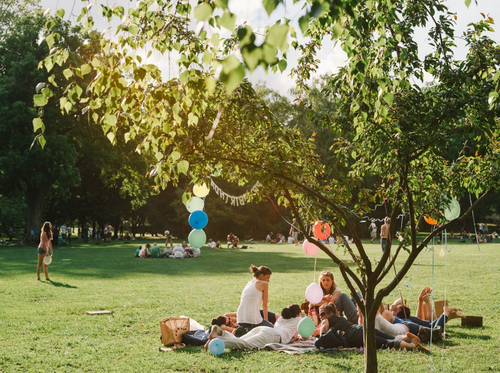 Group of people having a picnic under a tree in Margaret Island, Budapest, with colorful balloons and green grass surrounding them.
