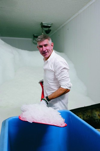 Eric Goeres stands in a freezing packing room at Alaska Ice Corp, holding a bag of ice above a blue bin.
