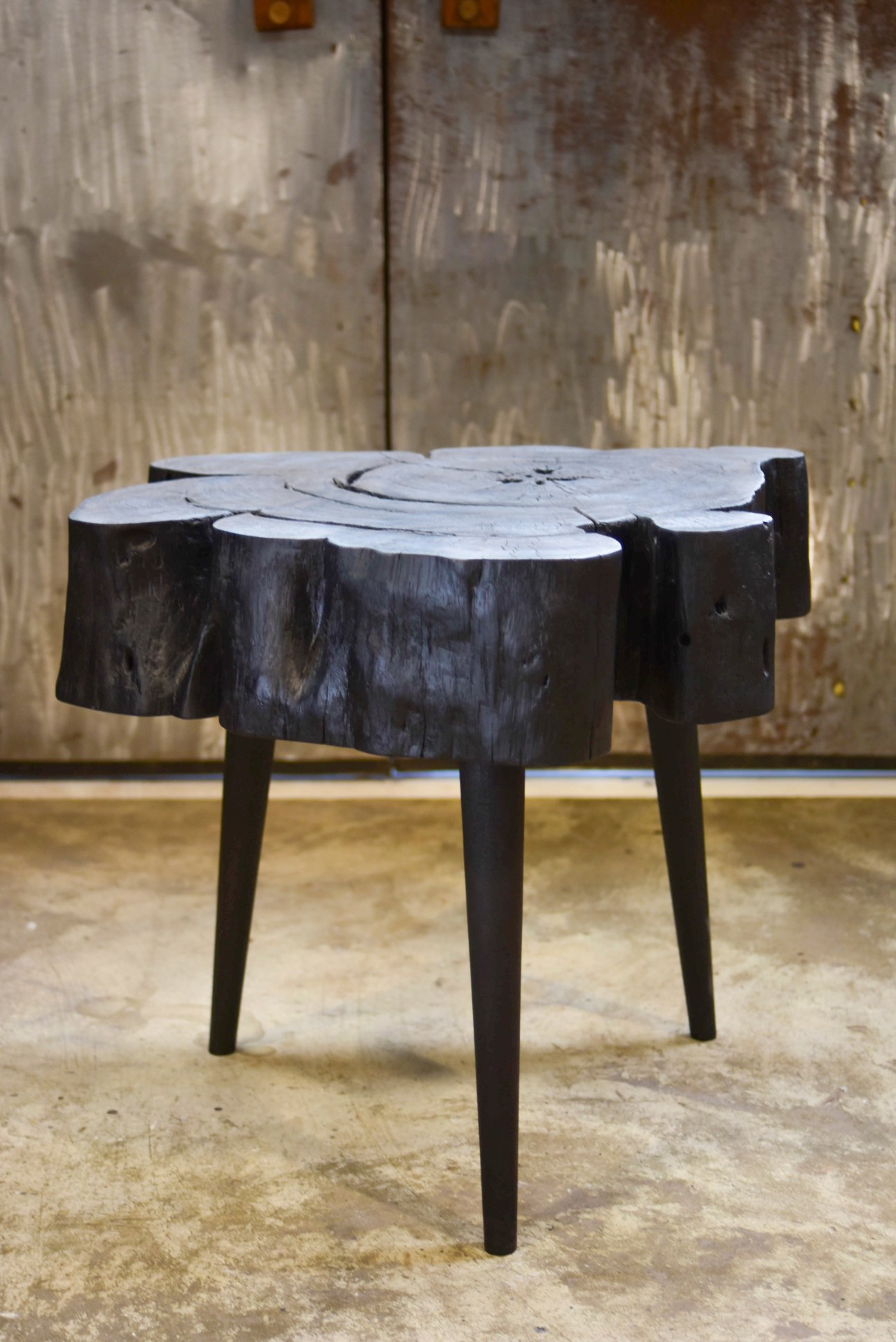 Dark wooden stool with organic top surface and tapered legs against weathered wall.