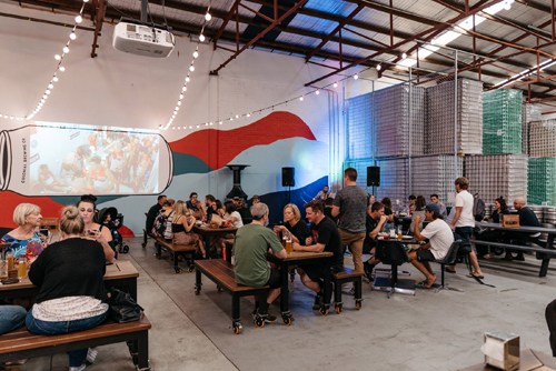 Interior of Colonial Brewing Company in Melbourne with patrons seated at communal tables beneath industrial ceiling and brewery floor visible.