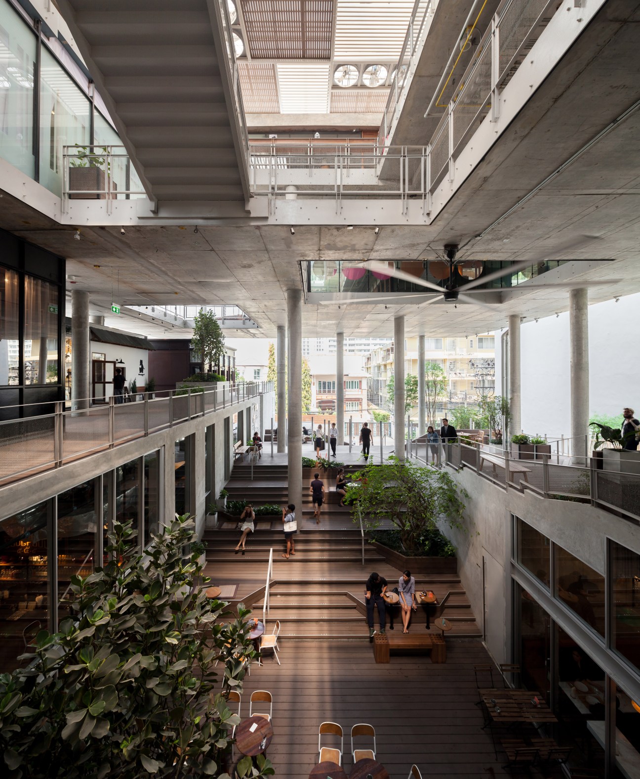 Wide wooden staircases at The Commons shopping mall in Bangkok with people sitting and walking through a multi-level atrium with plants and natural light.