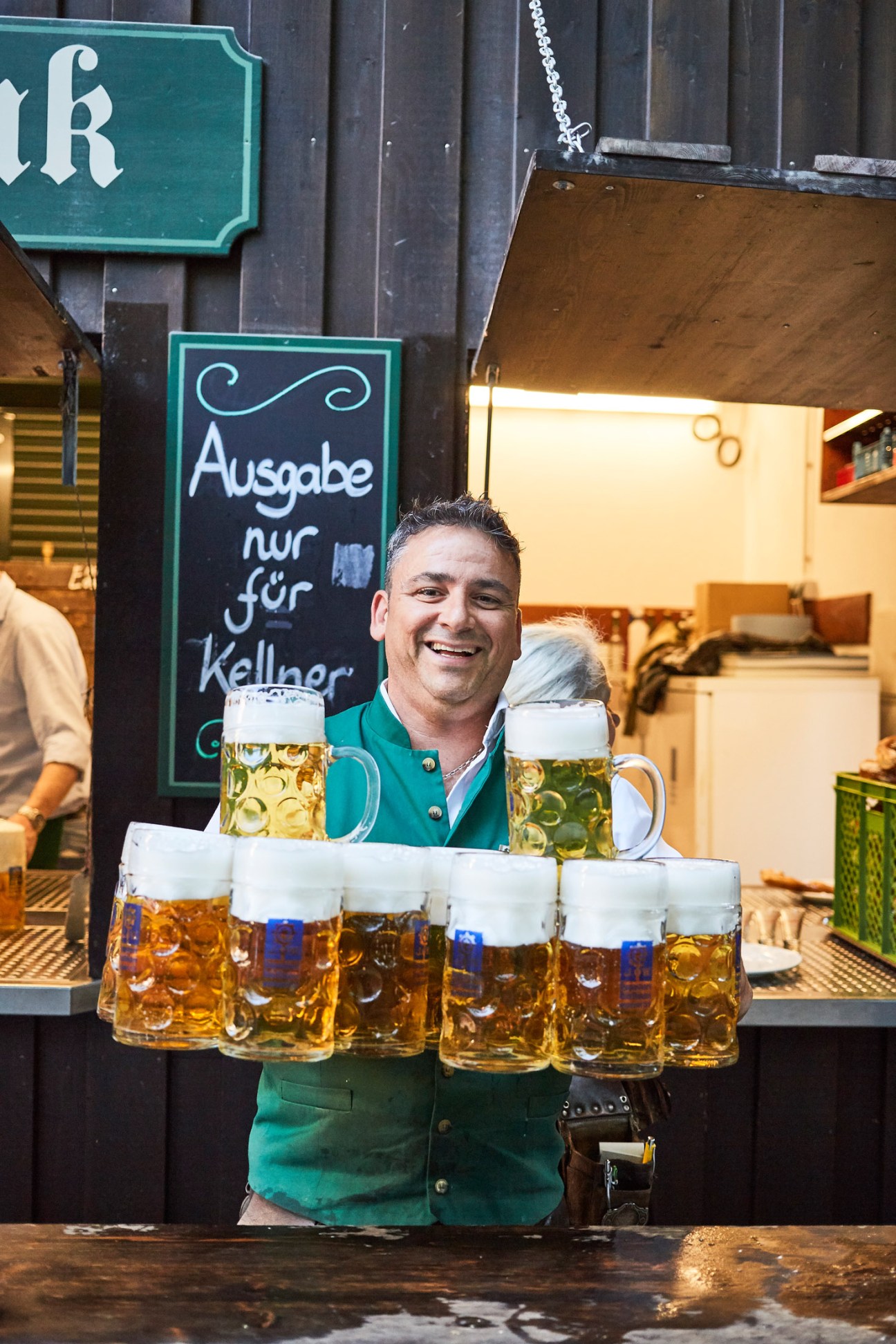 Bavarian beer hall server in traditional green vest holding multiple steins of beer in front of a chalkboard menu.