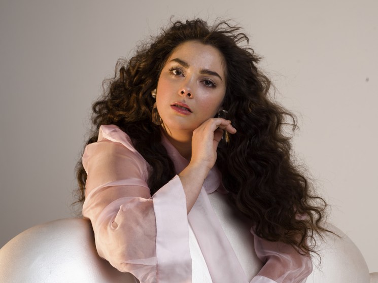 Woman with long dark wavy hair wearing a pink sheer shirt against a neutral background.