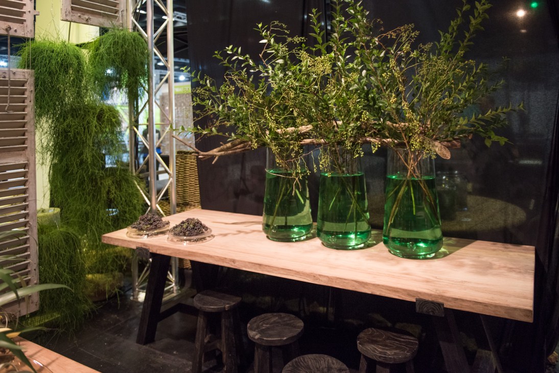 Trade fair display with wooden table, green glass vases with plants, and moss wall installation.
