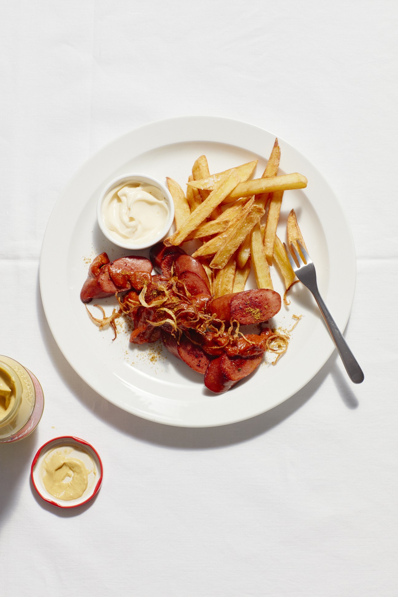 Plate of currywurst with golden chips, curry sauce, crispy onions, and mayonnaise on white plate.