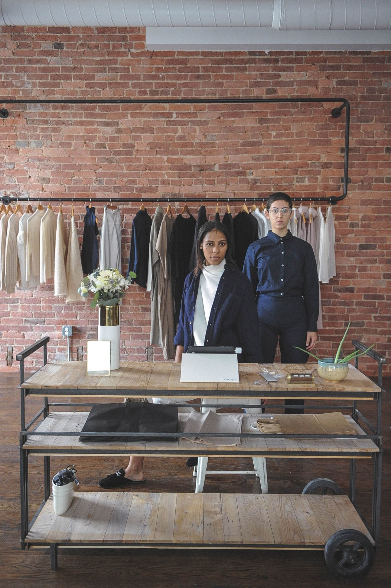 Two people stand behind a wooden counter in Shopkeepers store with exposed brick wall and clothing rack behind them.