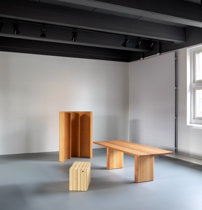 Wooden furniture display in gallery space with table, shelving unit, and stool.