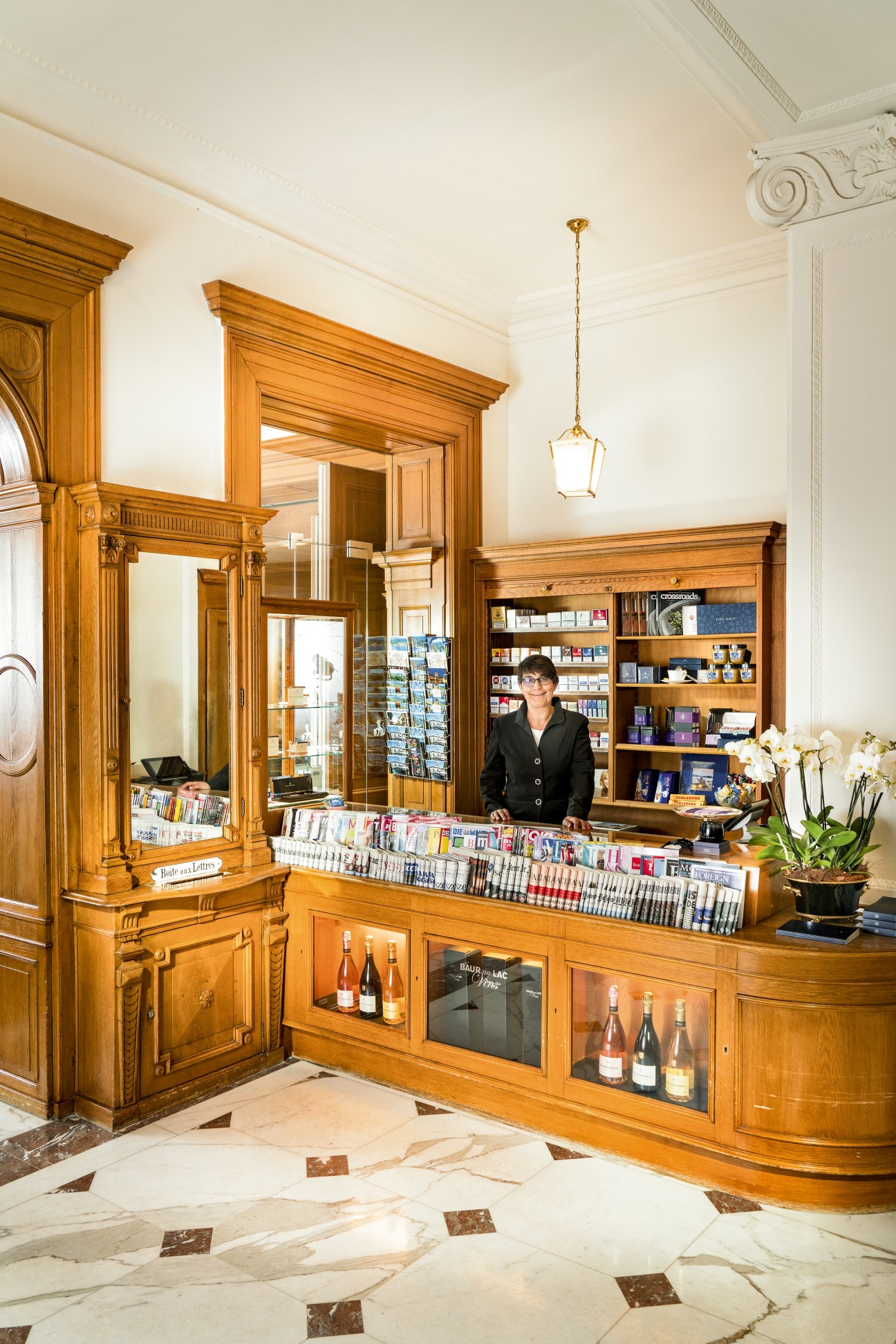 Woman standing behind a wooden kiosk counter displaying newspapers and magazines at Baur au Lac hotel in Zürich.