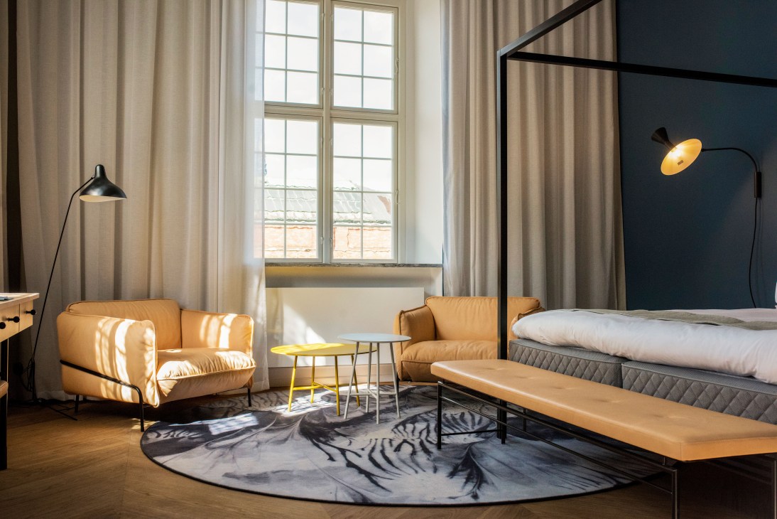 Modern hotel bedroom with tan leather armchair, black canopy bed, and curved wall sconce lamp.
