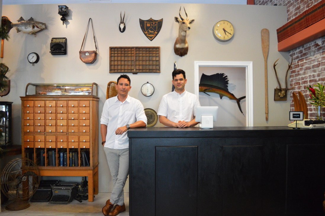 Two men in white shirts stand behind a counter in Hound and Quail shop surrounded by vintage curiosities and taxidermy.