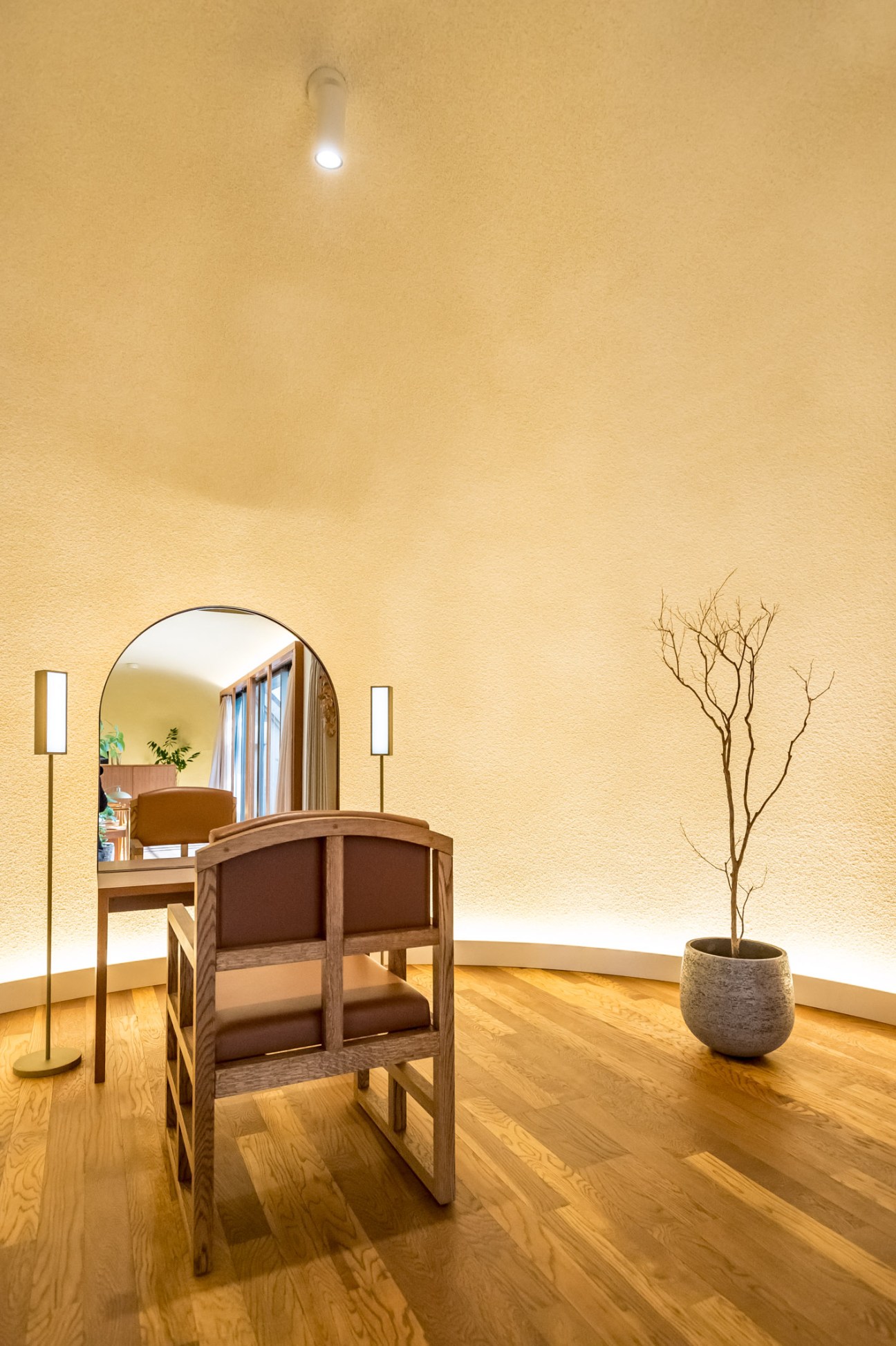 Minimalist styling room at Linda salon with wooden flooring, warm yellow walls, mirror, wooden chair with brown upholstery, brass floor lamp, and potted plant.