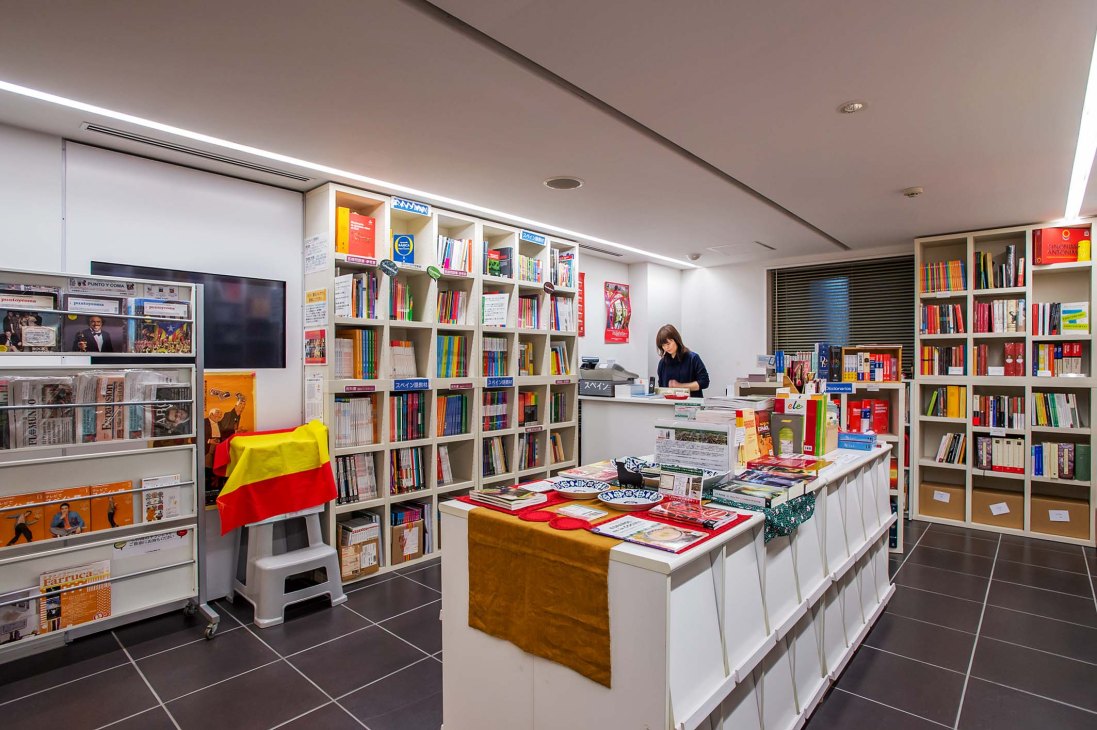 Library interior at Tokyo's Instituto Cervantes with Spanish flag, bookshelves, and staff member at desk.