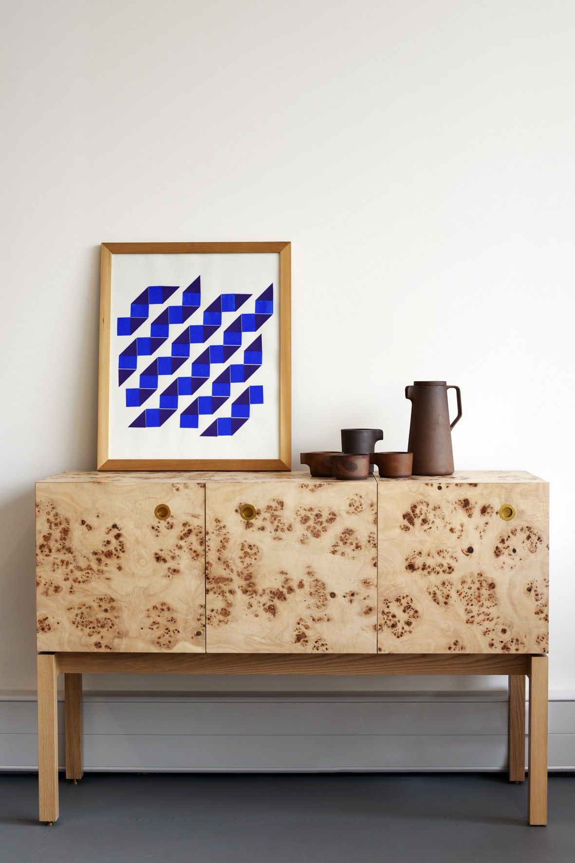 Wooden sideboard with burled veneer doors and ceramic vessels displayed on top next to framed geometric artwork.