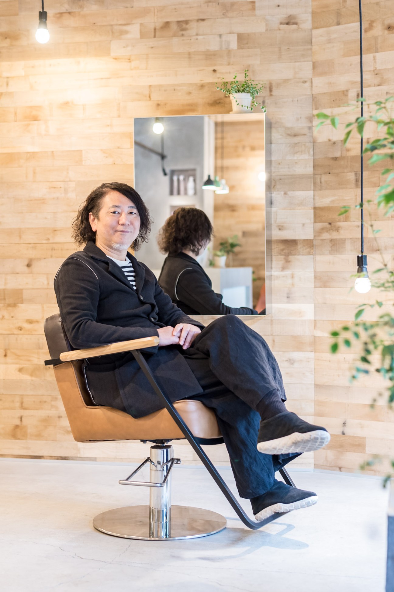 Architect Takeshi Okuwada sitting in a modern salon chair against a light wood wall with pendant lighting.