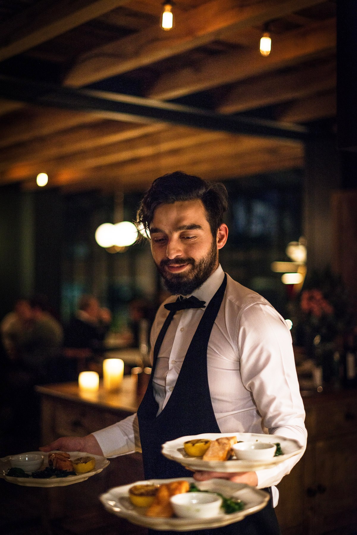 Male waiter in formal attire serving plated food in an elegant dining room with warm lighting.