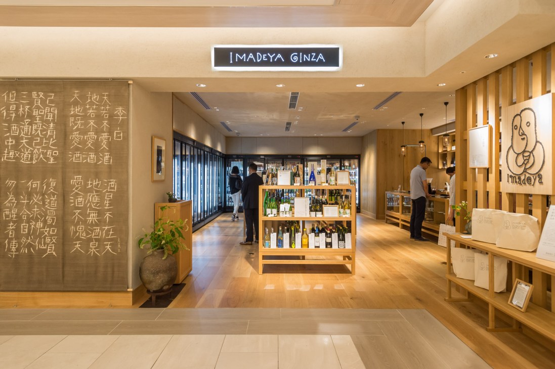 Interior of Imadeya Ginza liquor store with wooden shelving, signage, and customers browsing bottles.