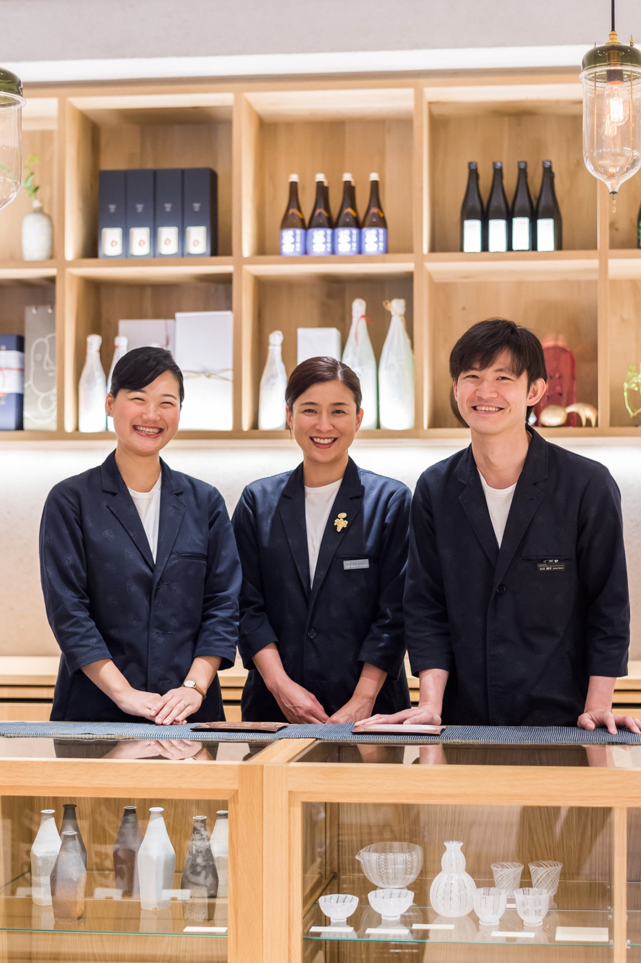 Three smiling staff members in navy blazers stand behind a wooden counter in a minimalist liquor shop with shelving displaying bottles and ceramic cups.