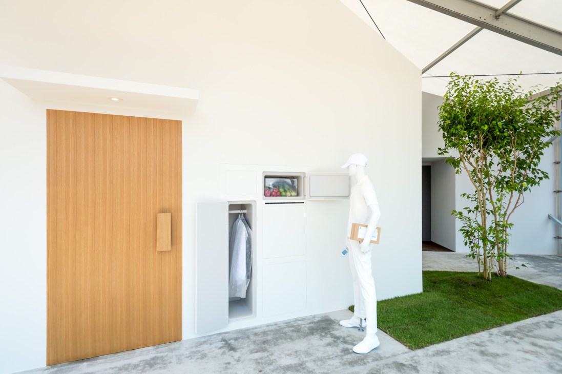 Modern minimalist interior with white walls, wooden door, external refrigerator access, white mannequin figure, and indoor plant under skylight.