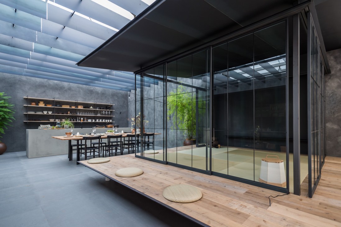 Modern minimalist interior with black-framed glass walls, wooden deck, dining table with black chairs, and open kitchen with shelving.