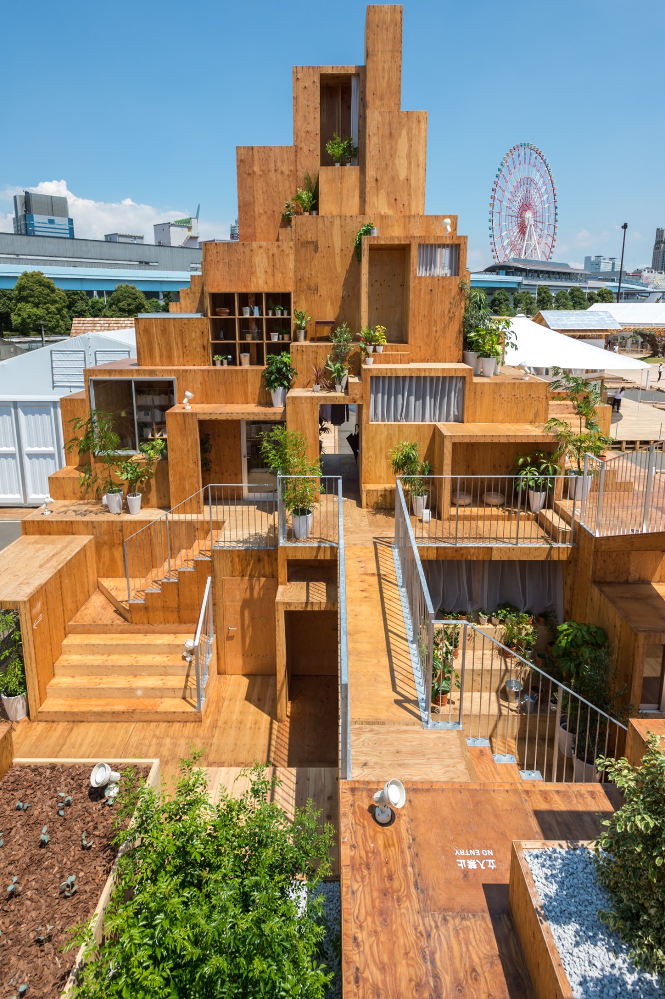 Wooden tower structure with stacked levels, open platforms, metal railings, and potted plants against blue sky and Ferris wheel.