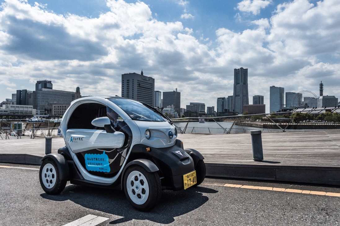 Nissan New Mobility Concept electric two-seater car parked on waterfront promenade with city skyline and river in background.