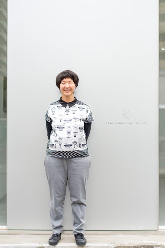 Woman with short dark hair wearing patterned sweater and grey pants standing against white gallery wall