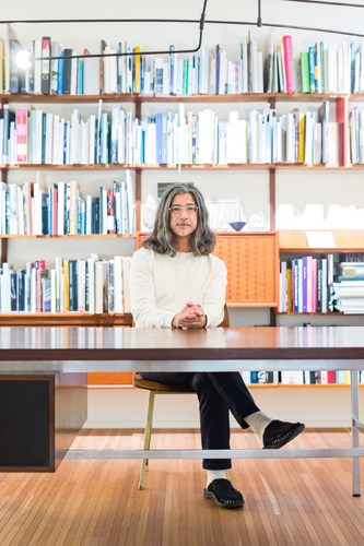 Woman with glasses and shoulder-length hair sits at wooden desk in front of floor-to-ceiling bookshelves