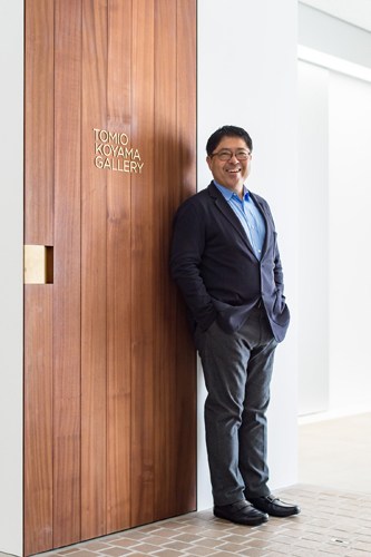 A smiling man in a dark suit stands next to wooden doors marked 'Tomio Koyama Gallery' in a modern white interior