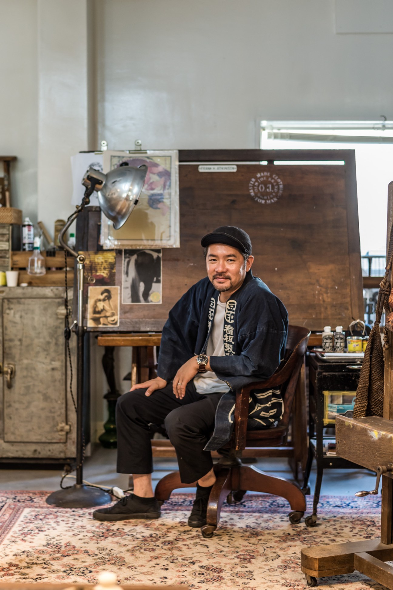 Man seated in vintage studio surrounded by antique furniture, industrial lamp, and framed photographs on dark wood wall.